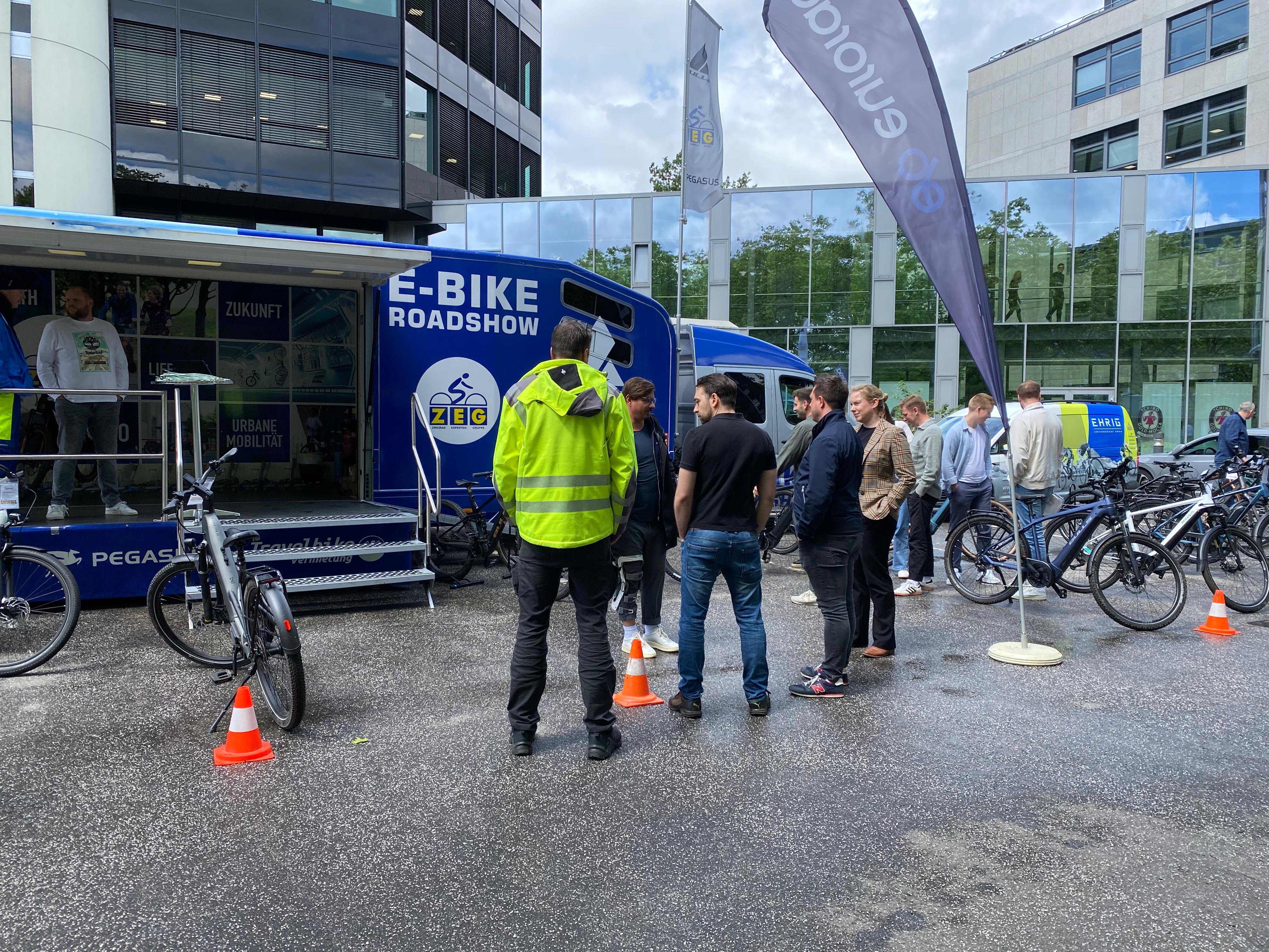 A group of people gathered around a blue van with 'E-BIKE ROADSHOW' written on it, various e-bikes on display, and a person in a high-visibility jacket near traffic cones.